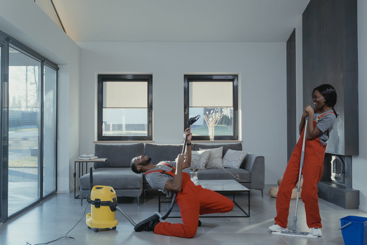 services-02 Fun and engaging cleaning session with a man and woman in overalls using cleaning tools in a modern living room.